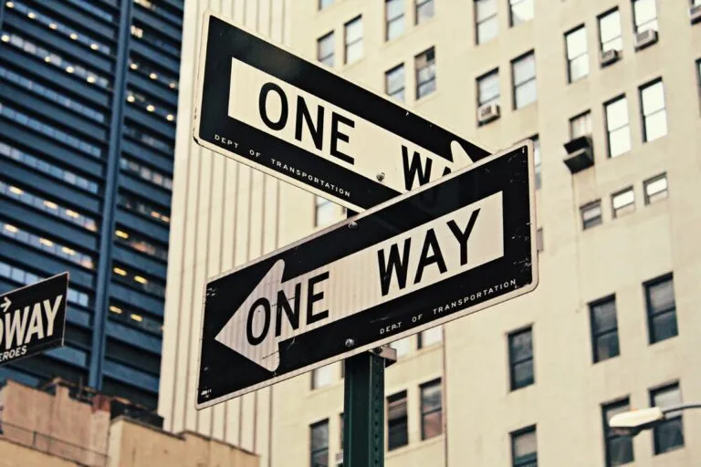 „Zwei Straßenschilder mit der Aufschrift ‚ONE WAY‘ zeigen in entgegengesetzte Richtungen vor einer Kulisse aus hohen Gebäuden.“