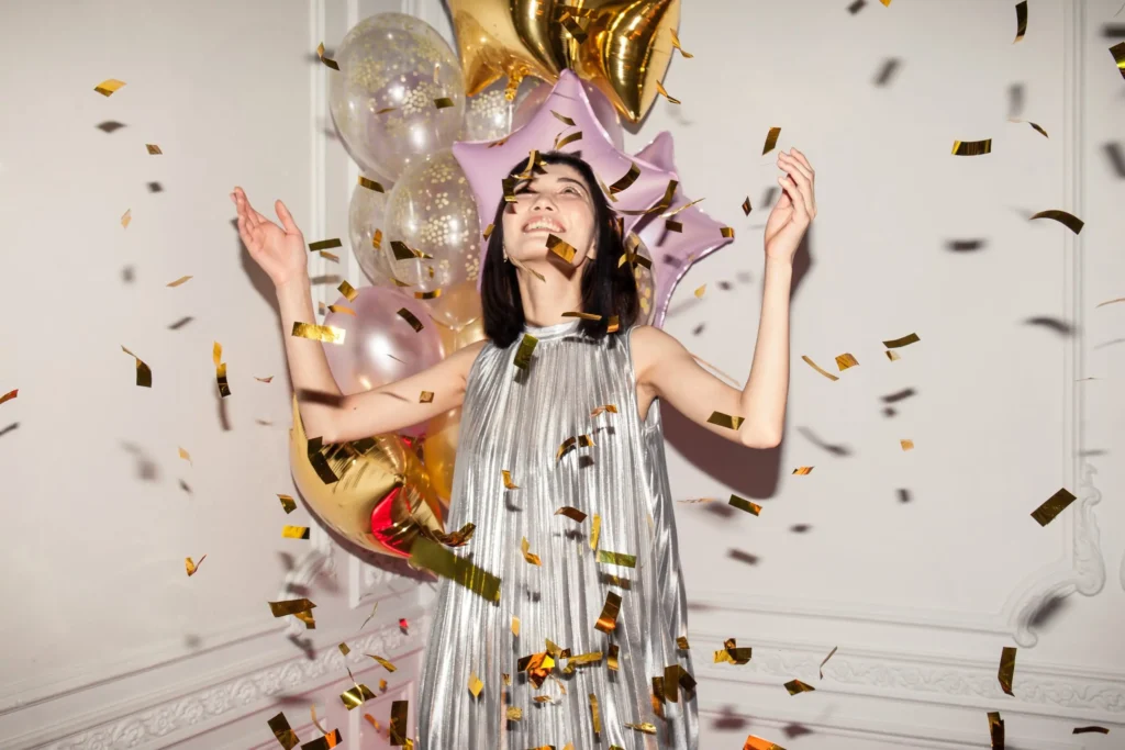 Person in silbernem Outfit steht vor Ballons und goldenen Sternen, während goldene Konfetti durch die Luft fliegen – festliche Stimmung