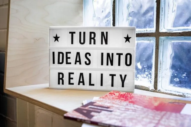 „Leuchtbox mit der Aufschrift ‚TURN IDEAS INTO REALITY‘ steht auf einem Holzregal neben Magazinen vor einem Glasbausteinfenster – Symbol für Innovation und Umsetzungskraft.“