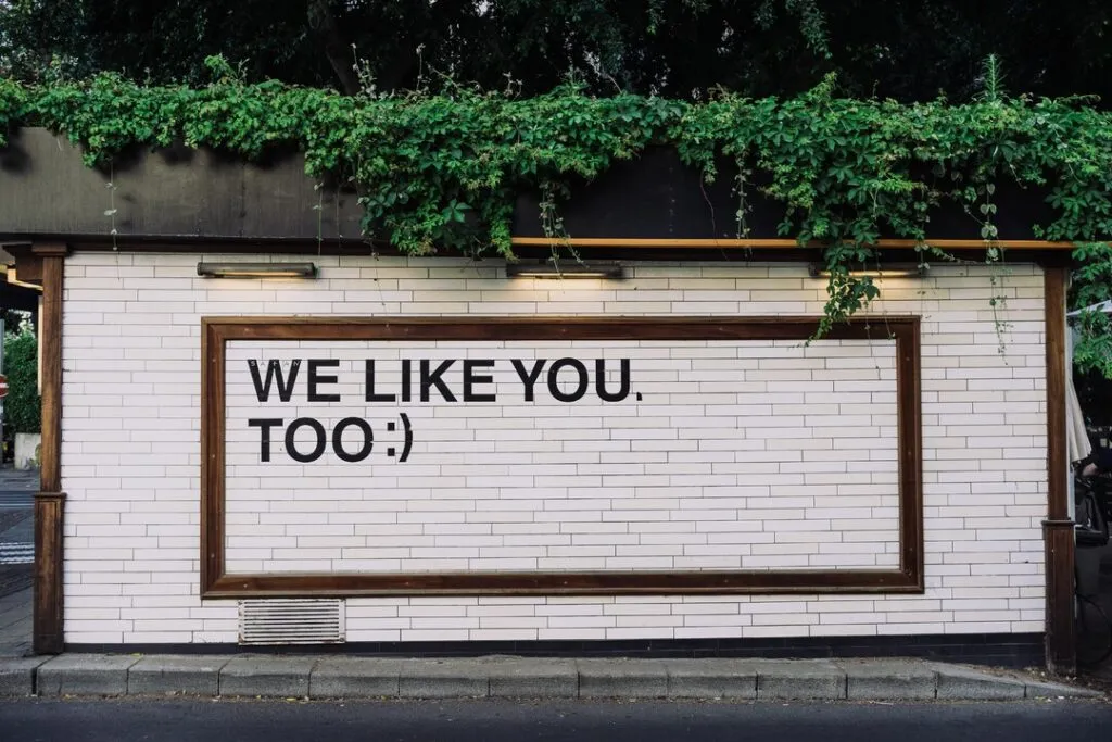 Weiße Wand mit schwarzer Aufschrift ‚WE LIKE YOU TOO :)‘, eingerahmt von Holz und umgeben von grünen Pflanzen – positive Botschaft im urbanen Umfeld