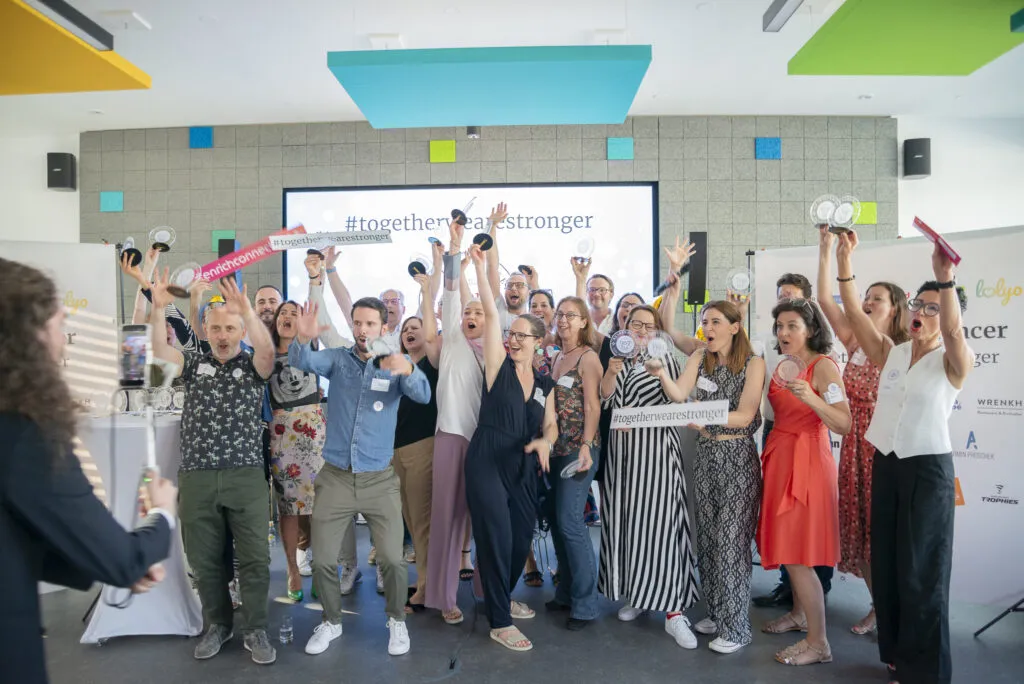 Gruppe von Menschen auf einer Bühne feiert mit Trophäen und Schildern vor einer Leinwand mit dem Hashtag #togetherwearestronger