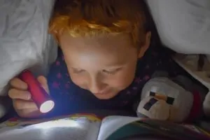 Kind liest unter einer Decke mit Taschenlampe und Buch – Symbol für Neugier und lebenslanges Lernen