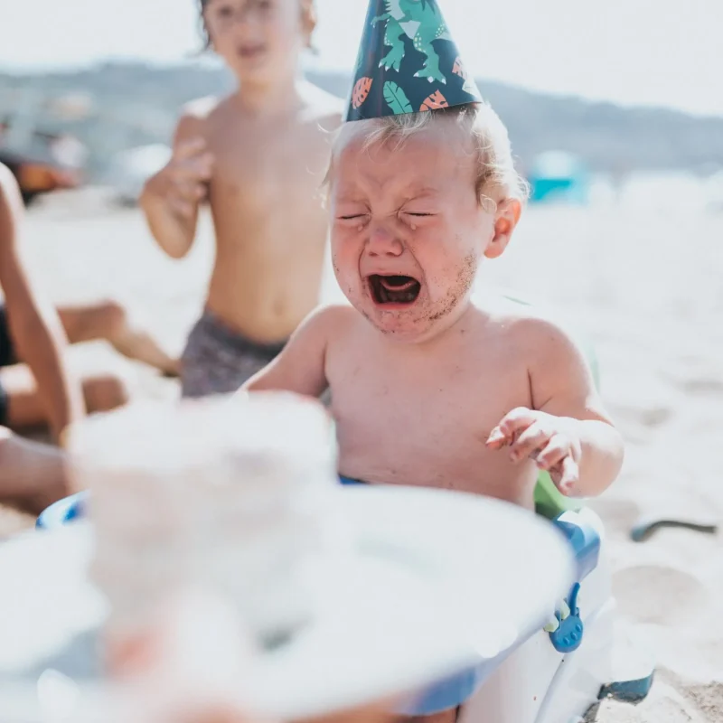 Kleinkind sitzt am Strand auf einem tragbaren Hochstuhl und trägt eine bunte Partyhut-Mütze, im Vordergrund ein Teller mit Kuchen.