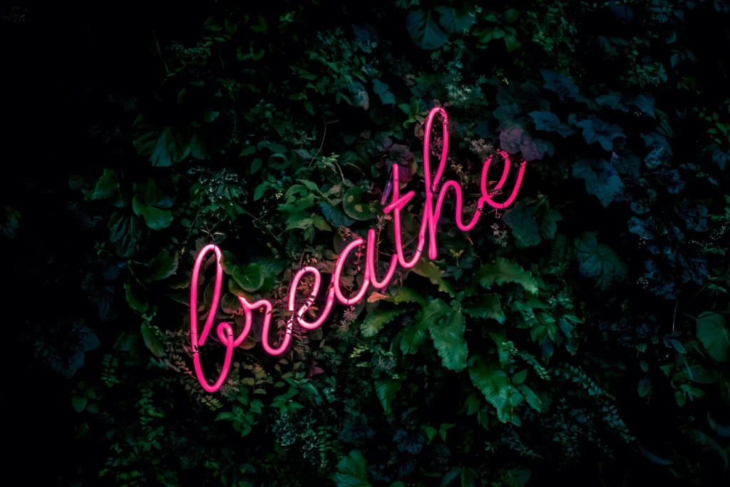Pinkes Neon-Wort ‚breathe‘ leuchtet vor einer grünen Pflanzenwand – Symbol für Achtsamkeit und Ruhe.