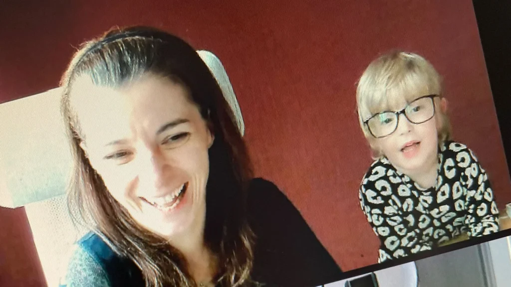 Laptop-Bildschirm zeigt ein virtuelles Meeting mit zwei Personen, eine sitzt auf einem Stuhl, die andere daneben, vor einer roten Wand.“