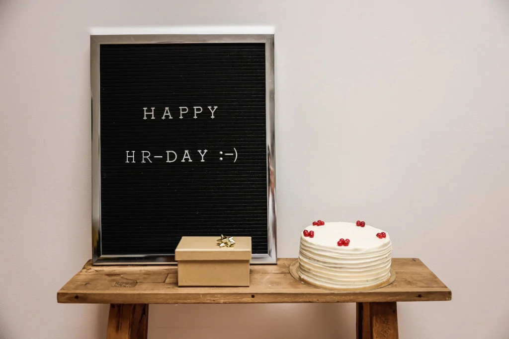 Holztisch mit einer weißen Torte, einer kleinen Geschenkbox und einem schwarzen Letterboard mit der Aufschrift ‚HAPPY HR-DAY :-)