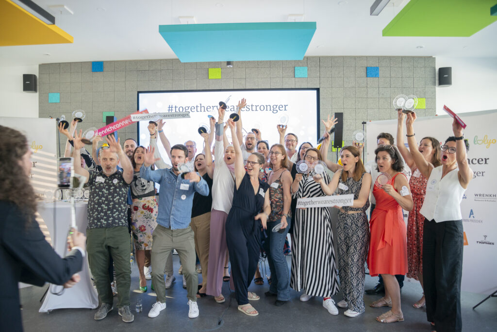 Gruppe von Menschen auf einer Bühne feiert mit Trophäen und Schildern vor einer Leinwand mit dem Hashtag #togetherwearestronger