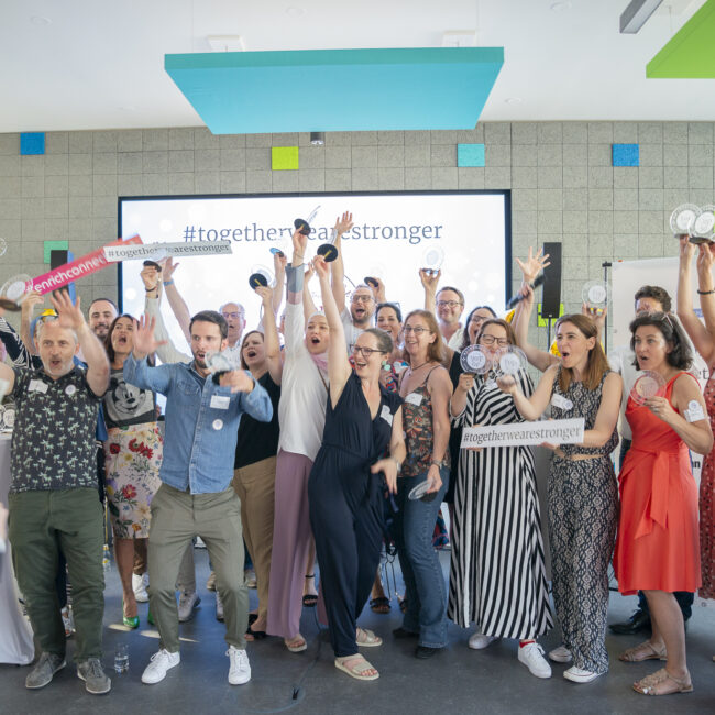 Gruppe von Menschen auf einer Bühne feiert mit Trophäen und Schildern vor einer Leinwand mit dem Hashtag #togetherwearestronger