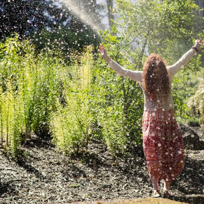 Person steht in einem sonnigen Garten mit ausgebreiteten Armen, während Wassertröpfchen in der Luft glitzern – Symbol für Befreiung und Klarheit